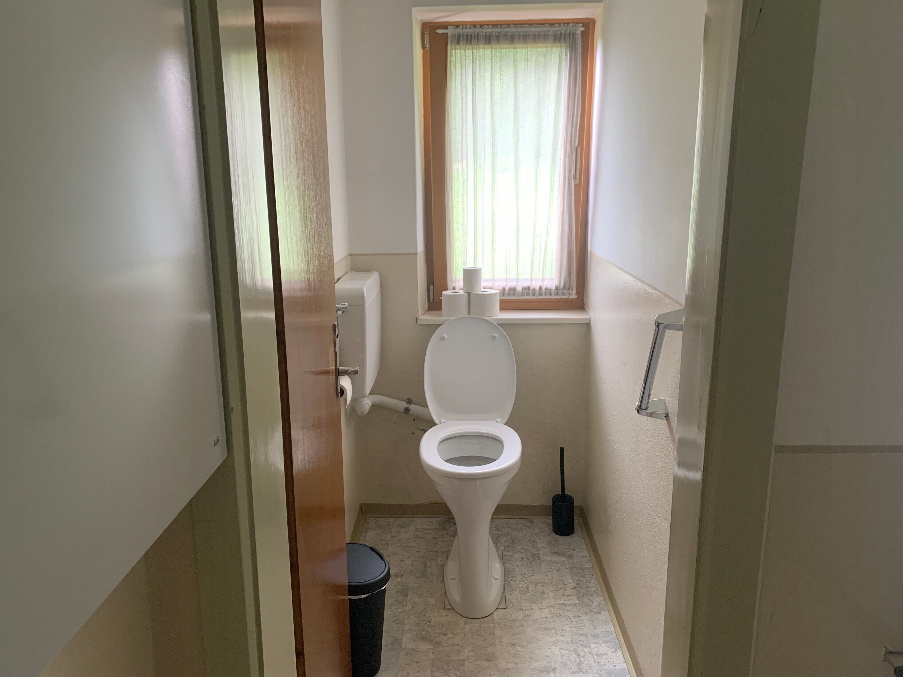 Ett litet, rent badrum med en toalett, ett fönster med gardin, en papperskorg och en toalettborste.