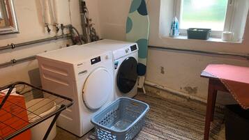 Ein Waschraum mit einer Frontlader-Waschmaschine, einem Trockner, einem Wäscheständer, einem Fenster mit Aussicht und einem kleinen Tisch mit Korb.