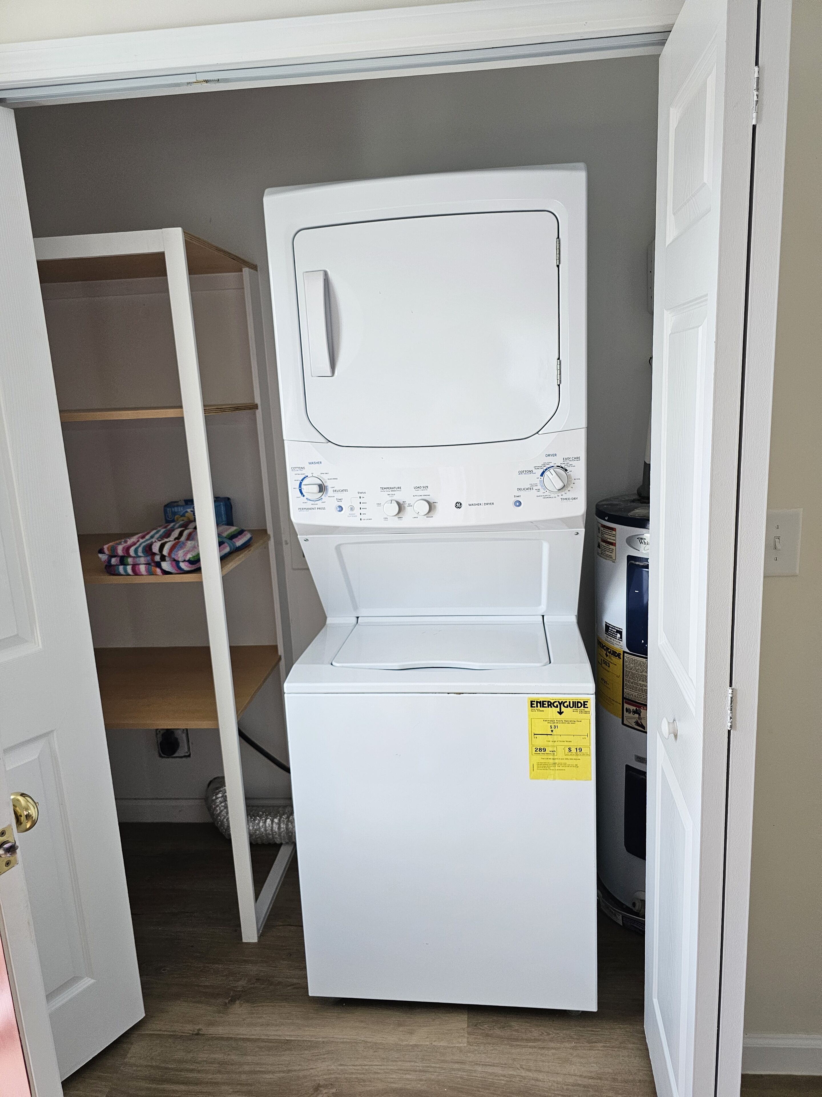 Une petite buanderie équipée d’un lave-linge et d’un sèche-linge à chargement frontal, d’une étagère en bois où sont pliés les vêtements, et d’une porte donnant sur une autre pièce.