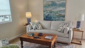 Un salon comprenant un canapé, une table basse et un fauteuil. On y trouve également un ventilateur de plafond, un tableau accroché au mur et une fenêtre avec des stores.