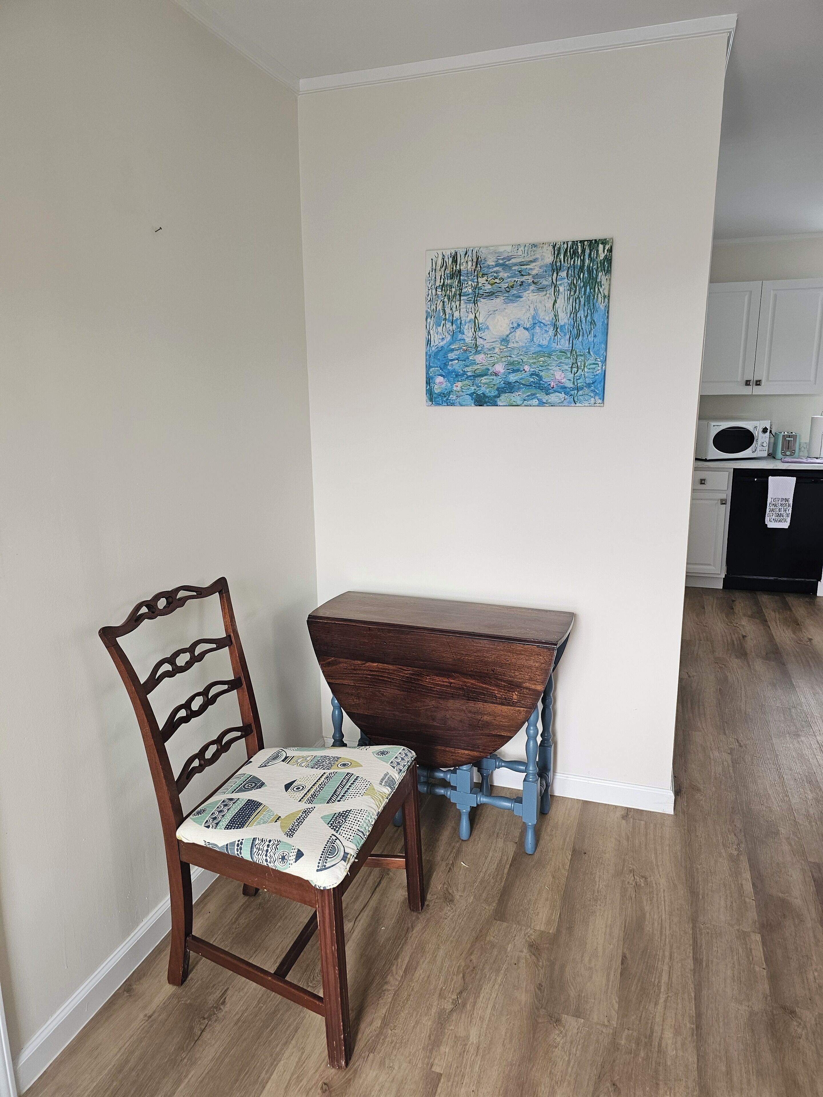 Een eetgedeelte met een houten tafel en stoel, een schilderij aan de muur, en een keuken op de achtergrond.