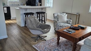 Un salon moderne avec un fauteuil en cuir, un fauteuil à motifs, une table basse en bois et un coin cuisine doté de meubles blancs et d’un réfrigérateur noir.