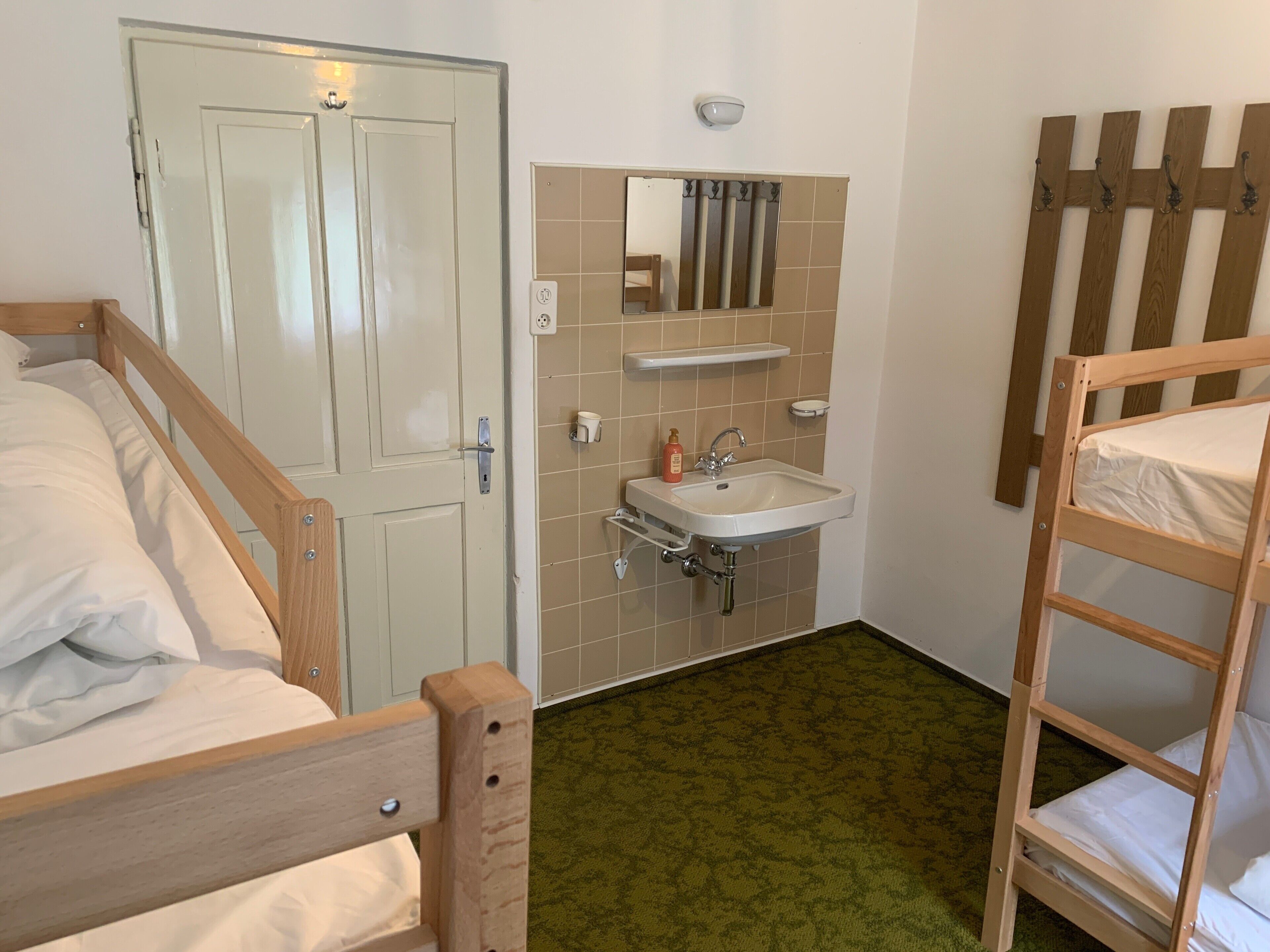 Quarto com uma cama beliche de madeira, uma cama de solteiro, banheiro com pia e espelho, e uma porta.