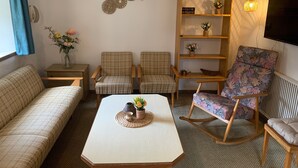 Un salon comprenant un canapé, deux fauteuils, une table basse agrémentée d’objets décoratifs, une bibliothèque en bois et un téléviseur fixé au mur.