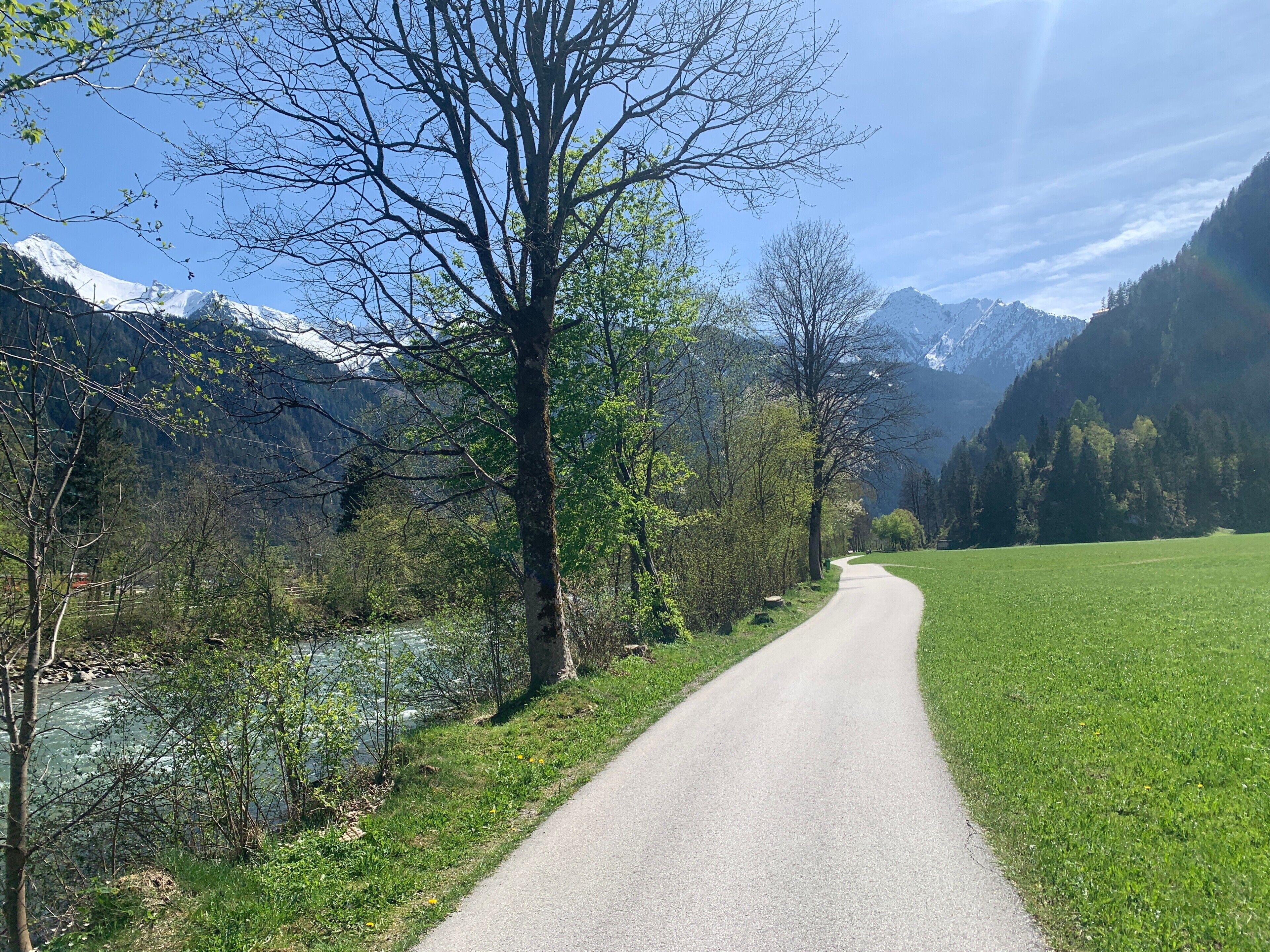 A paved path alongside a river, surrounded by trees and mountains.