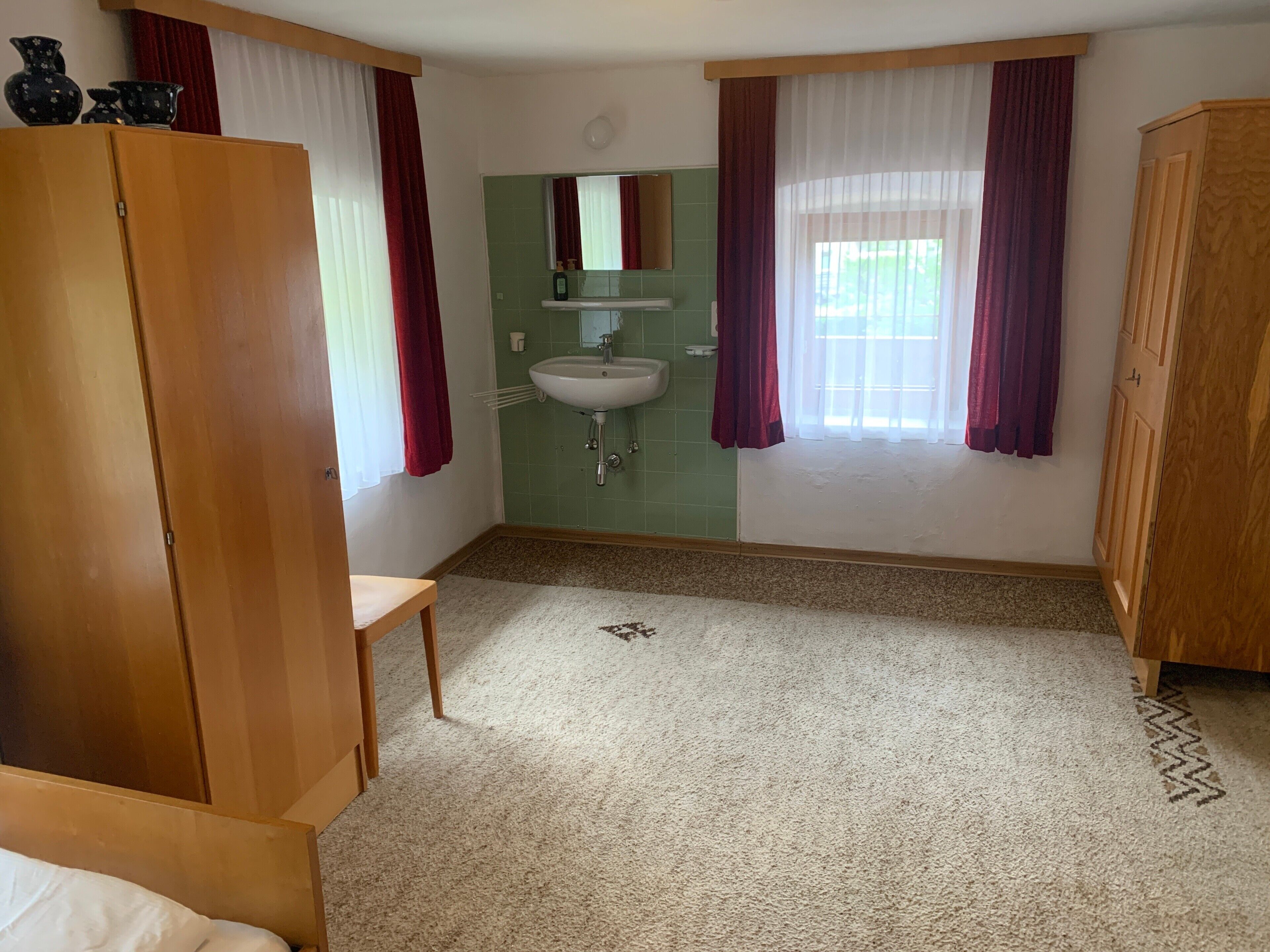Ein Schlafzimmer mit Holzschrank, einem kleinen Nachttisch, einem Einzelbett, einem Badezimmer mit Waschbecken und Spiegel sowie einem Fenster mit Vorhängen.