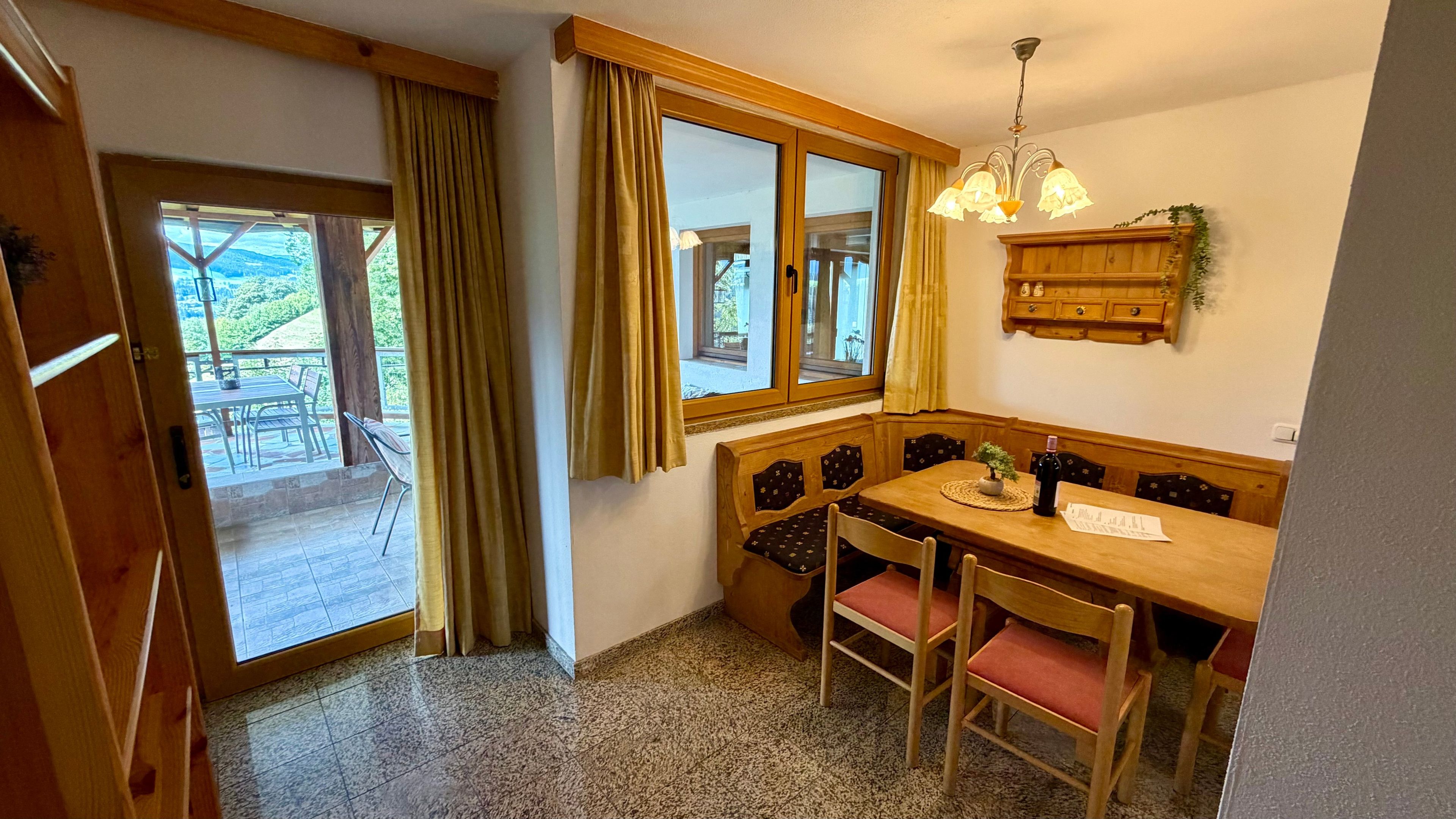 Un coin repas avec une table et des chaises en bois, un banc, une fenêtre avec des rideaux et un lustre.