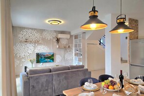 A modern dining area with a grey sofa, a dining table set with plates, glasses, and a fruit bowl. - Perla Holiday Villa 150, Lalzit Bay, Albania (Lalzit Bay)