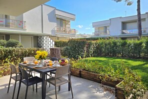 A modern outdoor dining area with a table set for breakfast, surrounded by a well-maintained garden and adjacent residential buildings. - Perla Holiday Villa 150, Lalzit Bay, Albania (Lalzit Bay)