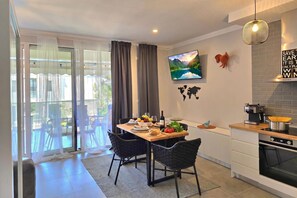 A modern kitchen with a dining area, a TV mounted on the wall, and a view of the outdoors through the sliding glass door.