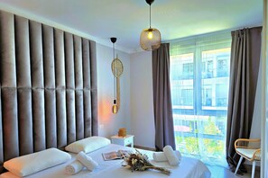 A modern hotel room with a large bed, a bedside table, a decorative wall hanging, a window with curtains, and a view of the outside.