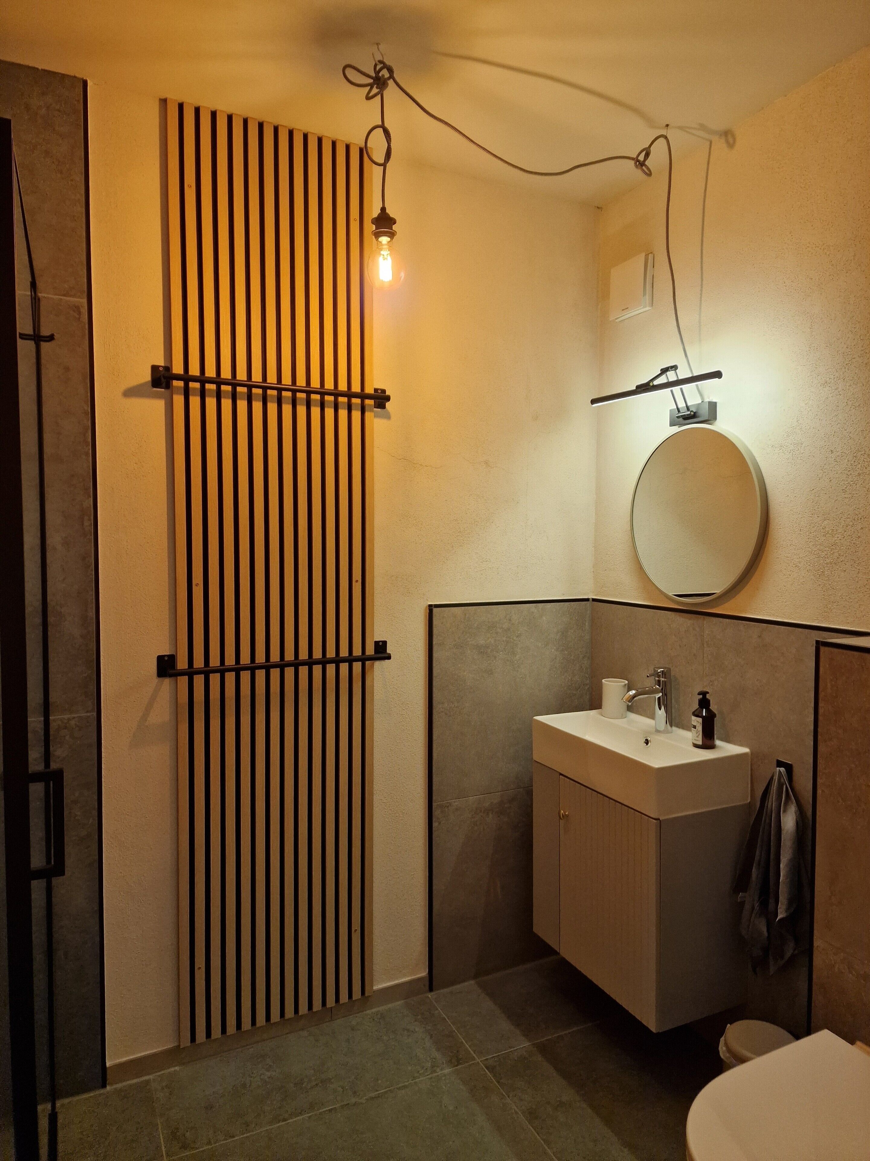 A bathroom with a modern design, featuring a vertical wooden slat door, a round mirror, a sink with a faucet, and a toilet.