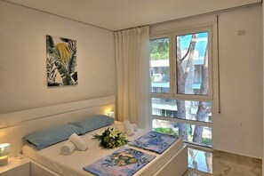 A hotel room with a bed, a window with curtains, a wall-mounted artwork, and a view of trees and buildings outside.