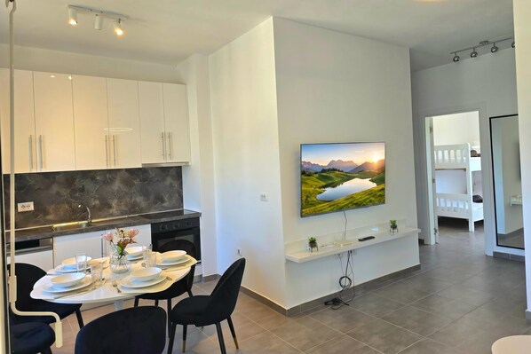 A modern kitchen with white cabinets, a dining area with a table set for four, and a mounted TV displaying a landscape.