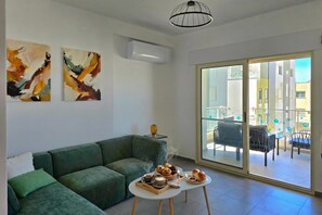 A modern living room with a green sofa, abstract wall art, a coffee table with pastries, and a balcony with outdoor furniture.