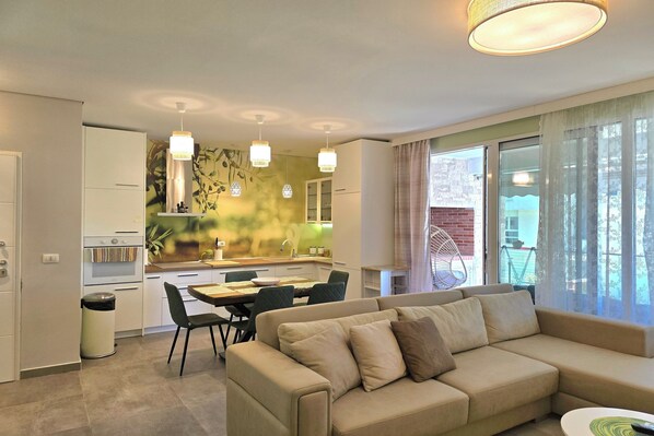 Eine moderne Wohnung mit Wohnzimmer, Essbereich und Küche. Es gibt ein beiges Sofa, einen Esstisch mit Stühlen und eine Küche mit weißen Schränken.