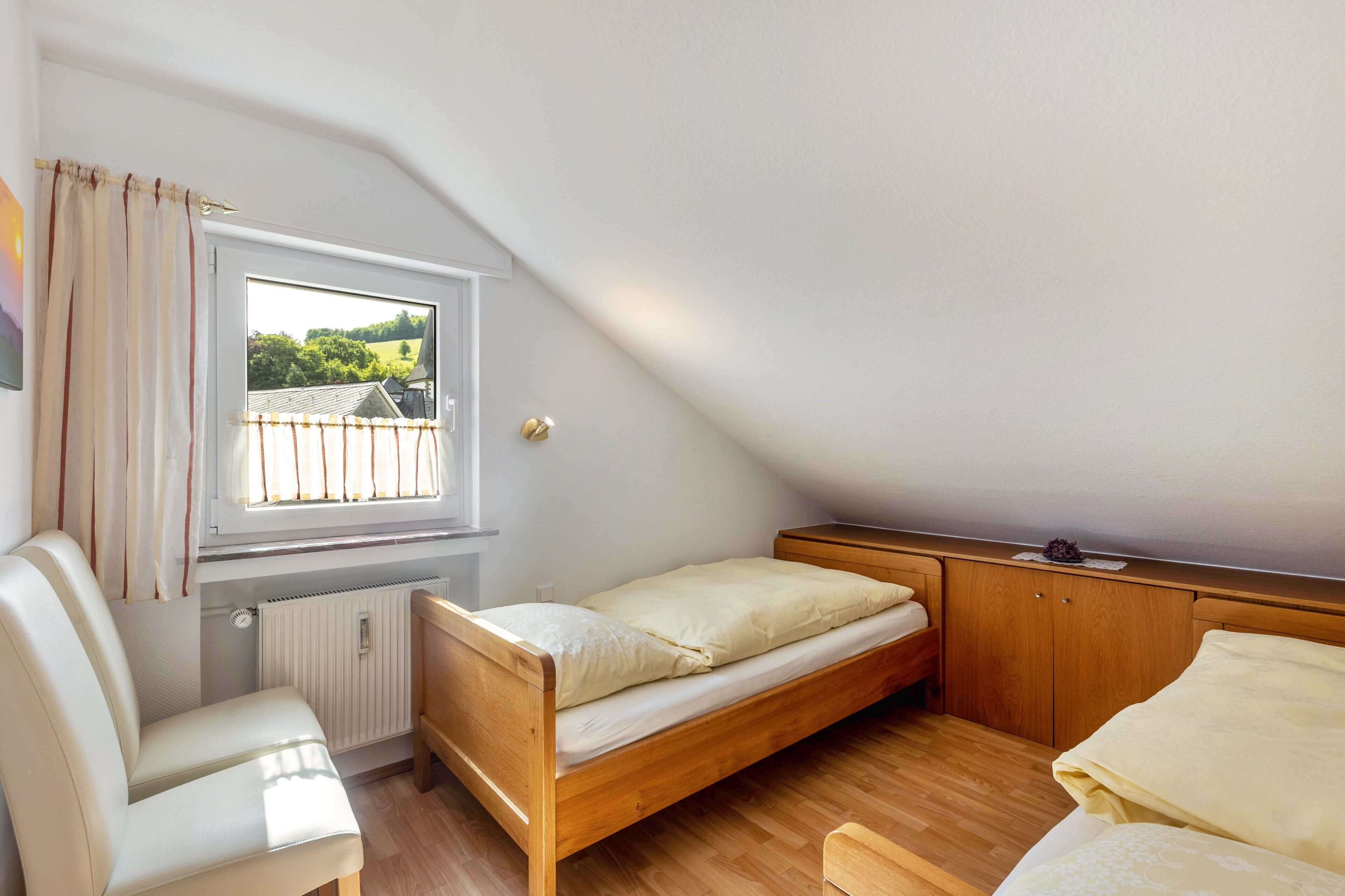 A small, single-room apartment with a sloped ceiling, a window with curtains, a radiator, a wooden bed, a wooden wardrobe, and a small table.
