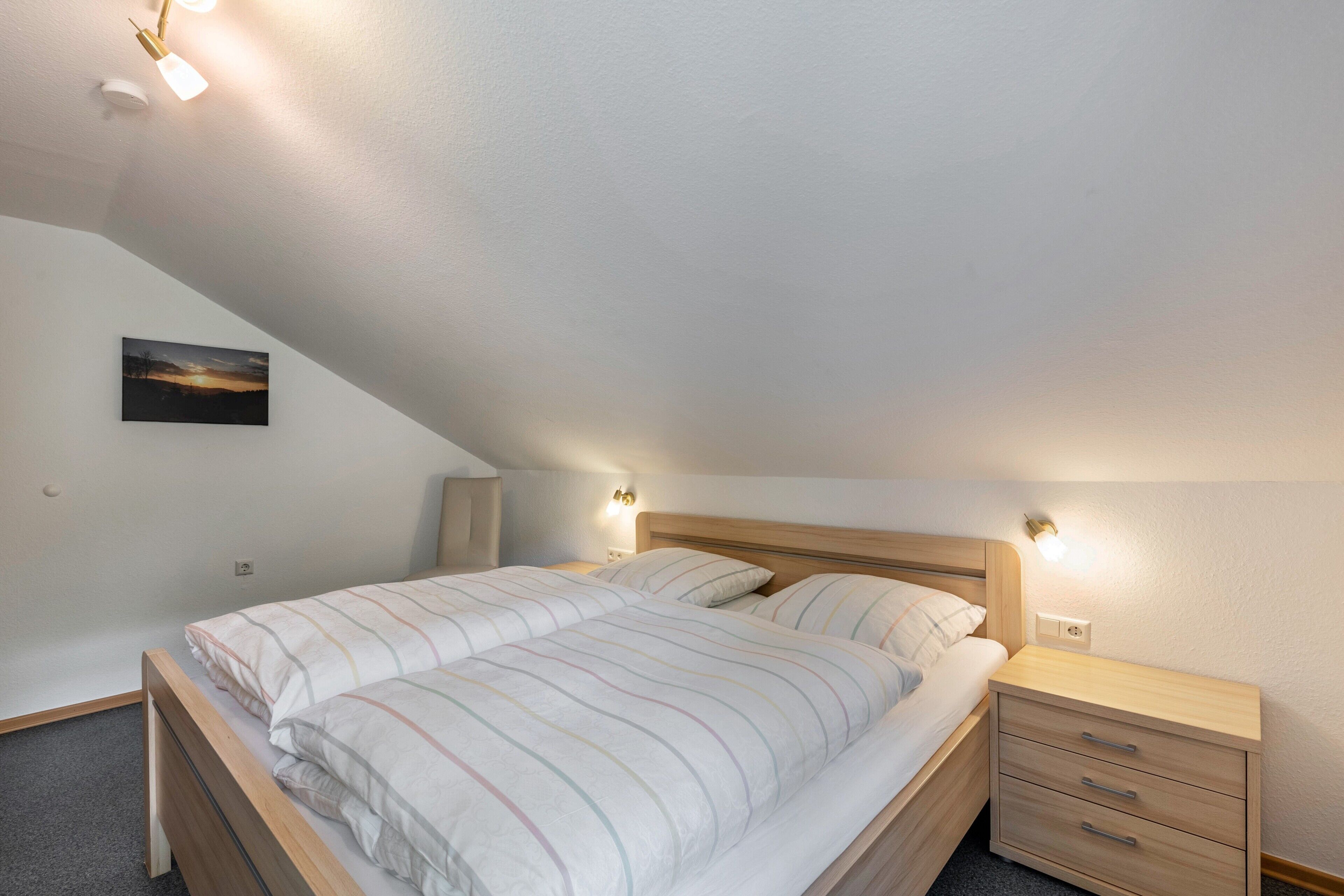 Ein Schlafzimmer mit schrägen Wänden, einem Bett mit gestreifter Bettwäsche, einem Nachttisch mit Lampe und einer Wandleuchte.