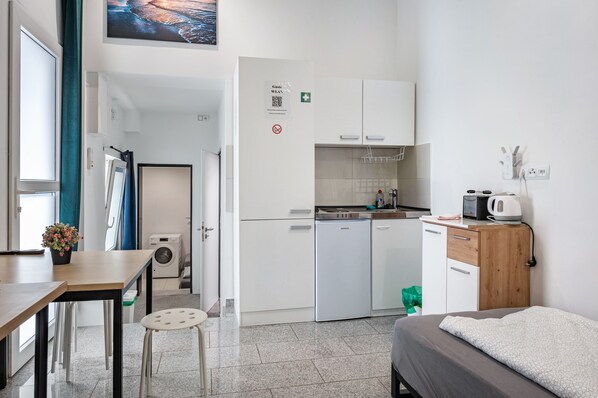 A compact hotel room with a kitchenette, a bed, a small table with a potted plant, and a view of the sea.