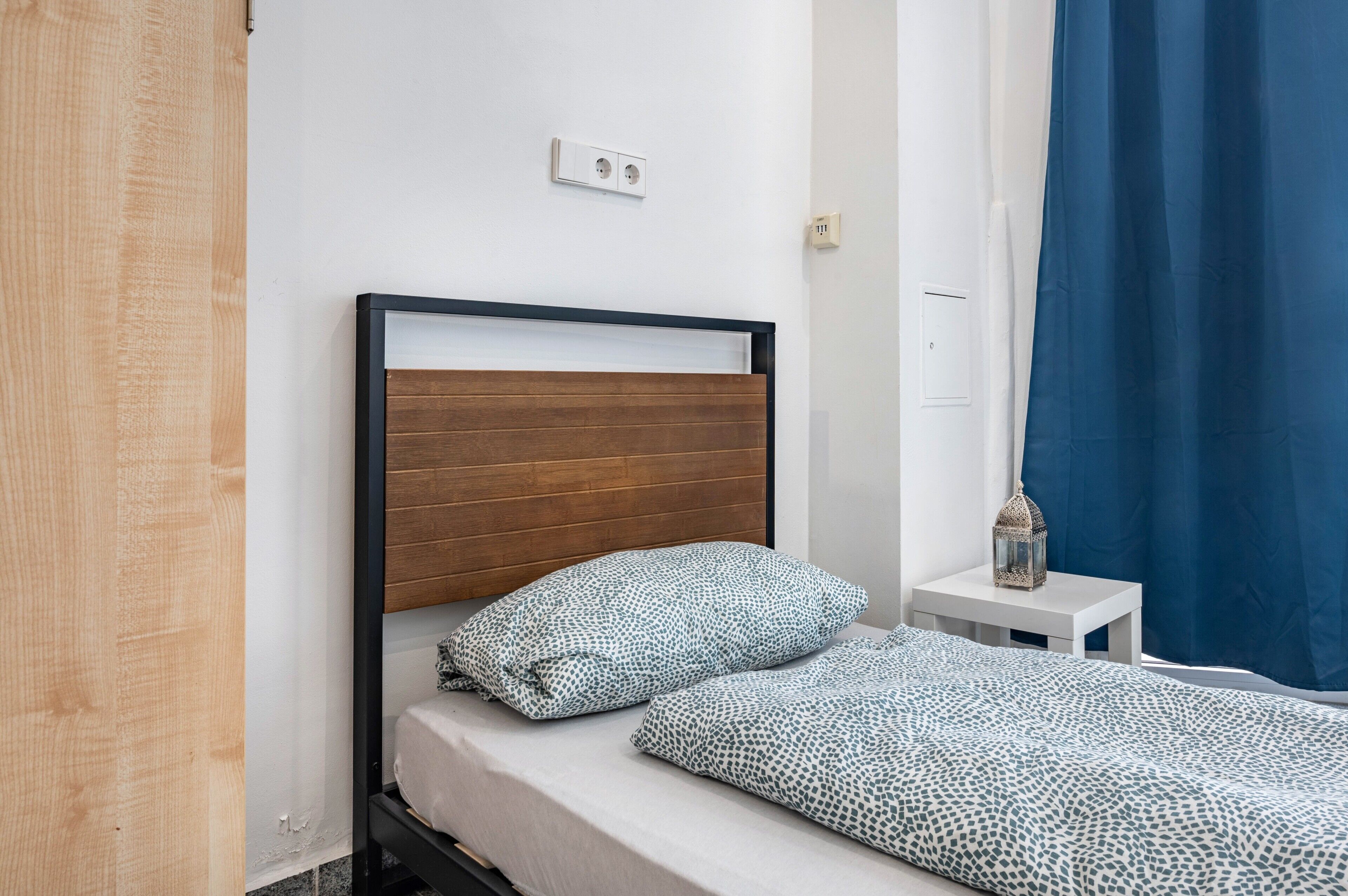 Un lit avec une tête de lit en bois, une table de chevet blanche surmontée d’une lanterne, et un rideau bleu.