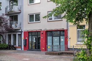 A building with a red door, a balcony with a metal railing, and a tree in the foreground.