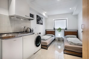 A compact hotel room with a kitchenette, a washing machine, a bed with a headboard, a bedside table with a plant, and a window with blinds.