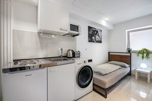 Une pièce compacte comprenant un lave-linge, un petit coin cuisine, un lit et un téléviseur fixé au mur.