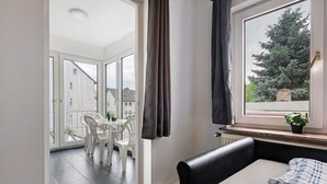 A room with a bed, a window with curtains, a balcony with a table and chairs, and a view of trees and buildings.