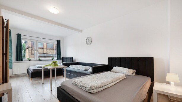 Quarto moderno com uma cama grande, um sofá-cama, uma mesinha com uma planta e uma janela com cortinas.