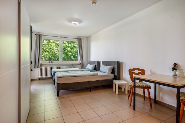 A bedroom with a bed, a small table, a chair, a window with curtains, and a radiator.