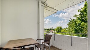 Un balcón con mesa y sillas, una pared blanca y vistas a zonas verdes y edificios.