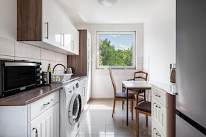 A compact kitchen with a washing machine, a microwave, and a window with a view of greenery.