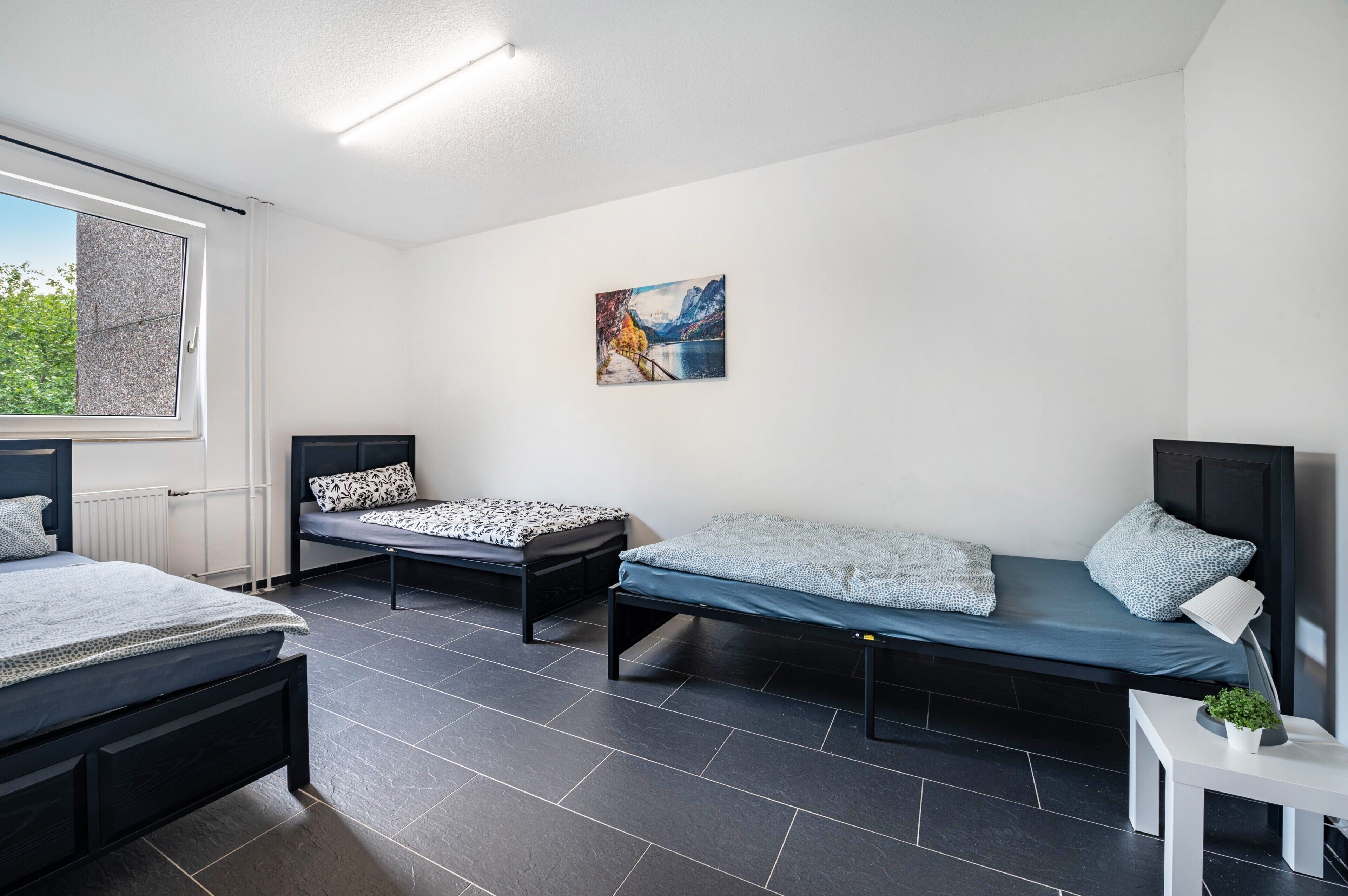 A room with two single beds, a small table with a plant, a painting on the wall, and a window with curtains.