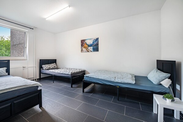 A room with two single beds, a small table with a plant, a painting on the wall, and a window with curtains.