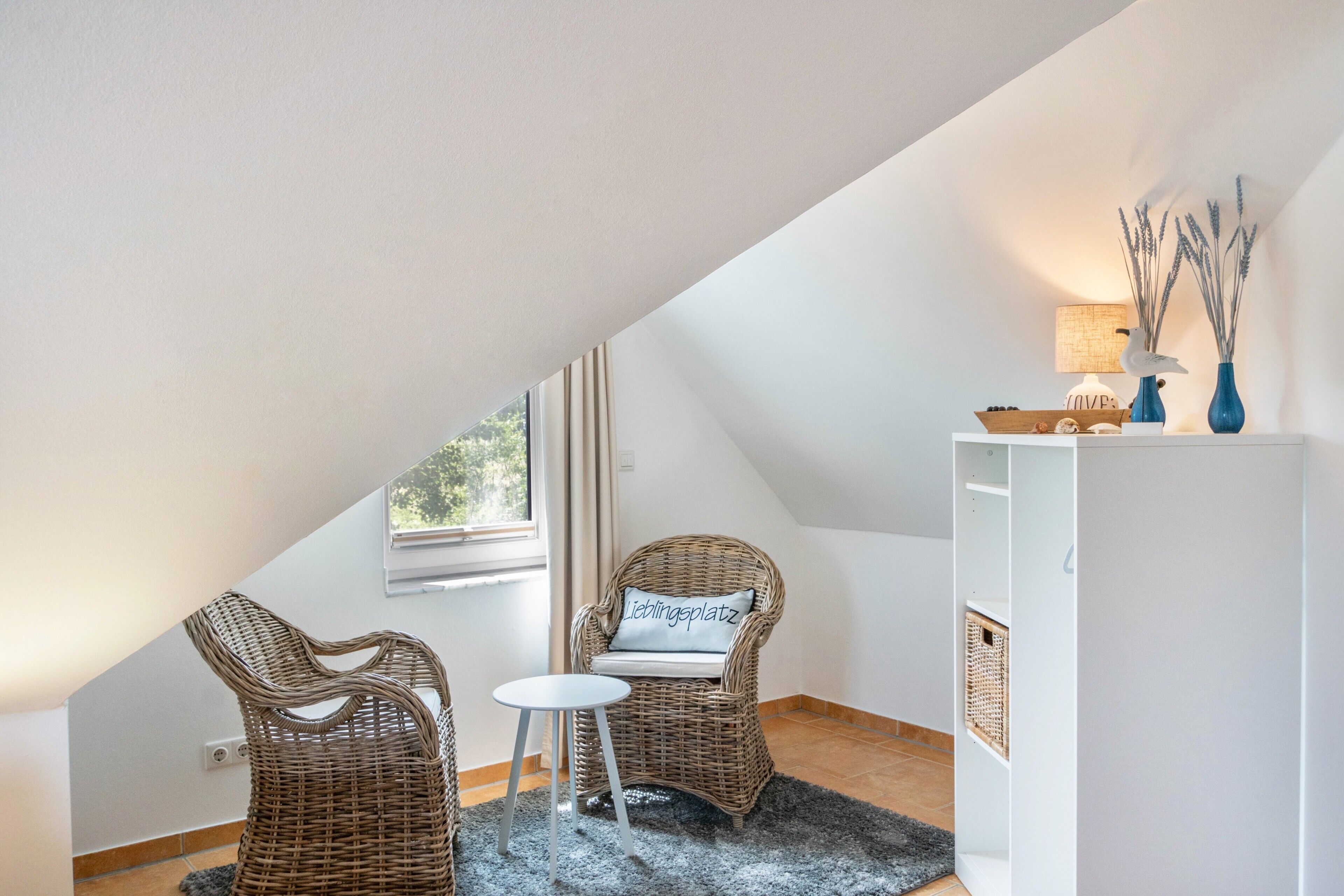 A room with a sloped ceiling, two wicker chairs, a small round table, a white cabinet, and a window with curtains.