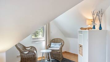 A room with a sloped ceiling, two wicker chairs, a small round table, a white cabinet, and a window with curtains.