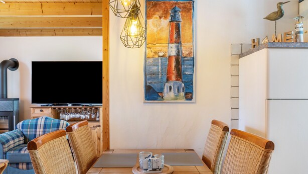 A dining area with a wooden table, wicker chairs, a painting of a lighthouse, and a television on the wall. - Holiday Home 'Hafenblick' with Private Terrace, Private Garden and Wi-Fi (Walchum)
