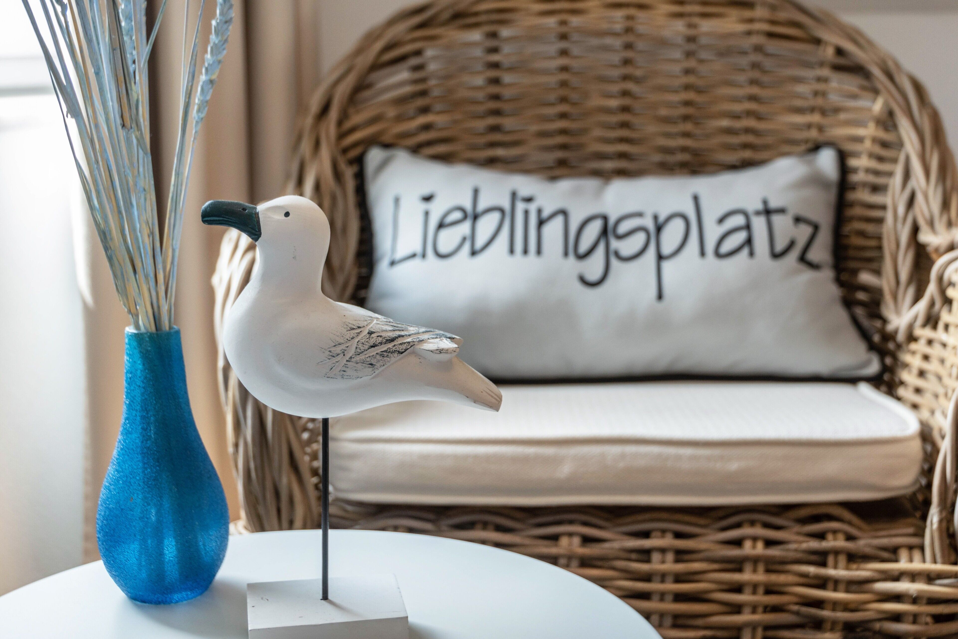 Ein Korbstuhl mit Kissen, beschriftet mit 'Lieblingsplatz', und eine weiße Vogelfigur auf einem weißen Tisch.