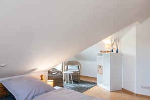 Quarto com cama, cadeira de vime, mesa pequena, armário branco e prateleira com objetos decorativos.
