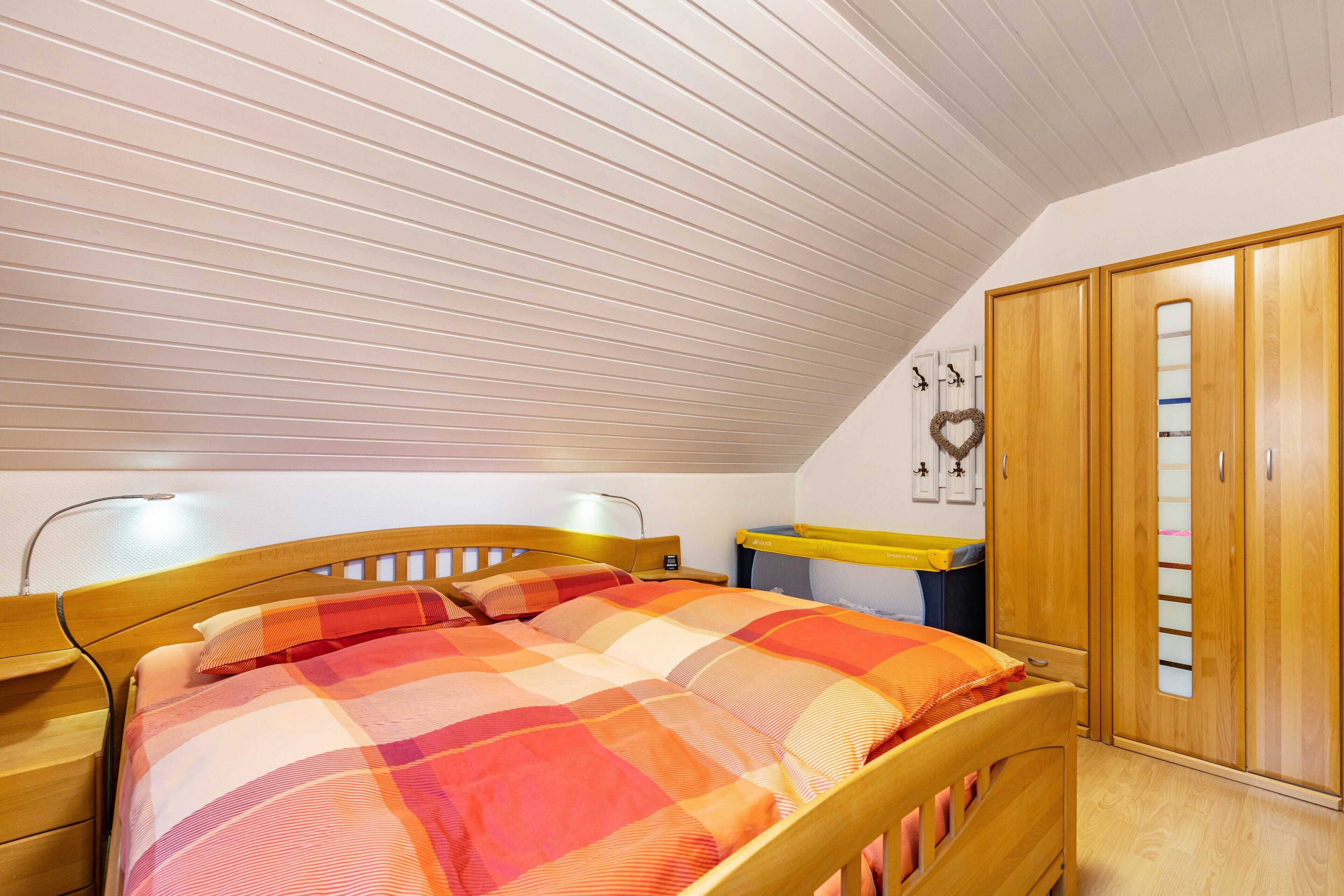 Une chambre avec un plafond en pente, un lit recouvert d’une couverture à carreaux, une armoire en bois et une table de chevet avec une lampe.