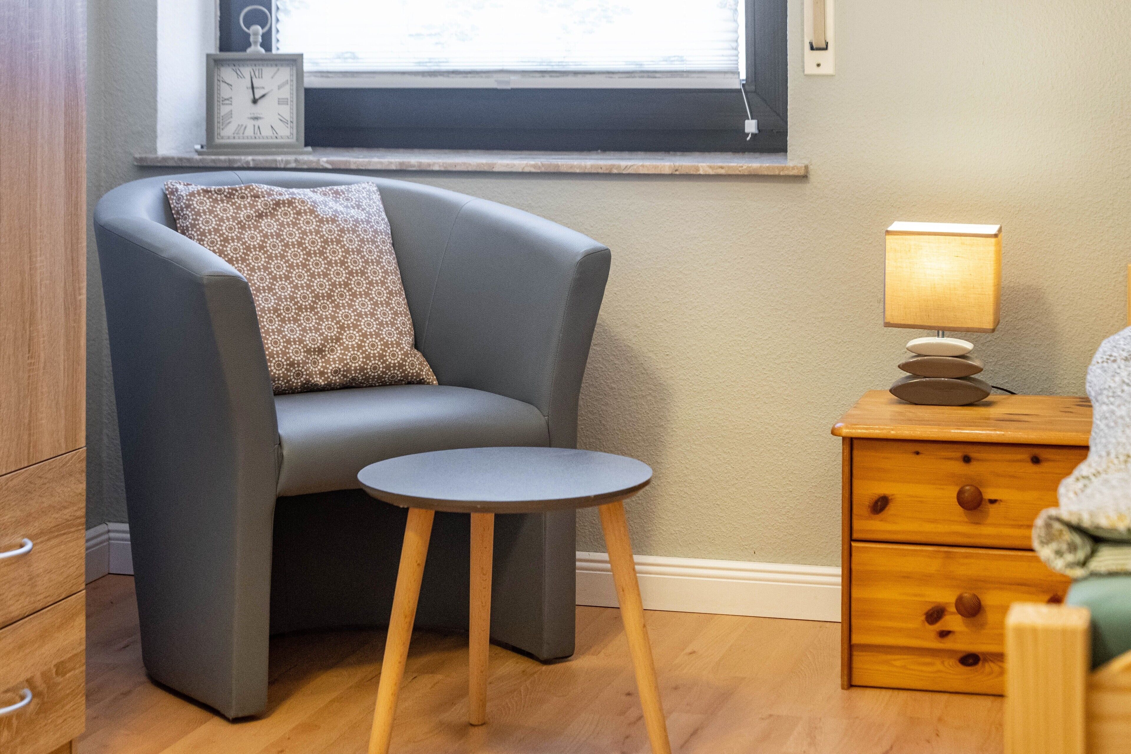 Une pièce d’angle avec un fauteuil gris, une petite table ronde, une table de chevet en bois avec une lampe et une horloge au mur.