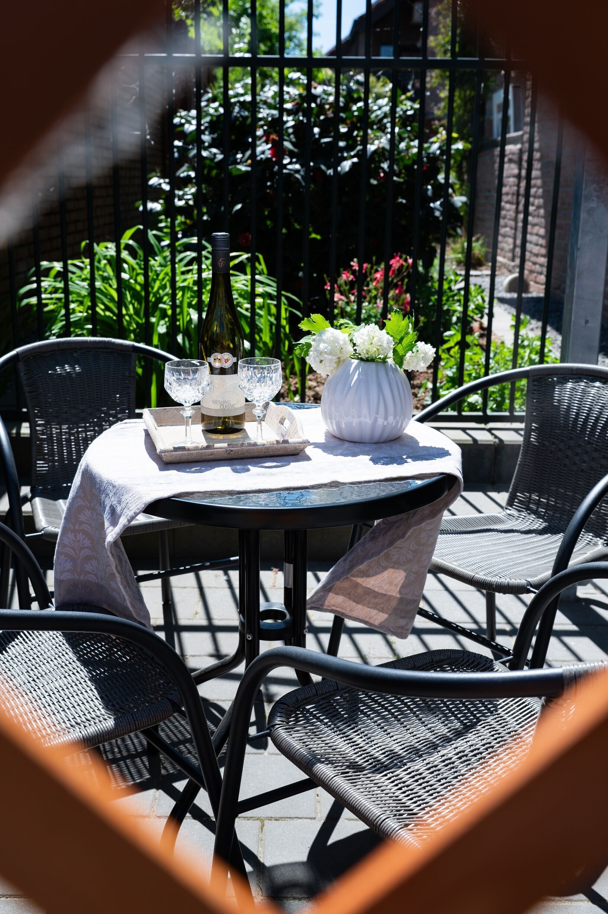 Un patio al aire libre con una mesa redonda preparada para dos personas, una botella de vino Riesling Classic, dos copas de vino y un jarrón con flores blancas.