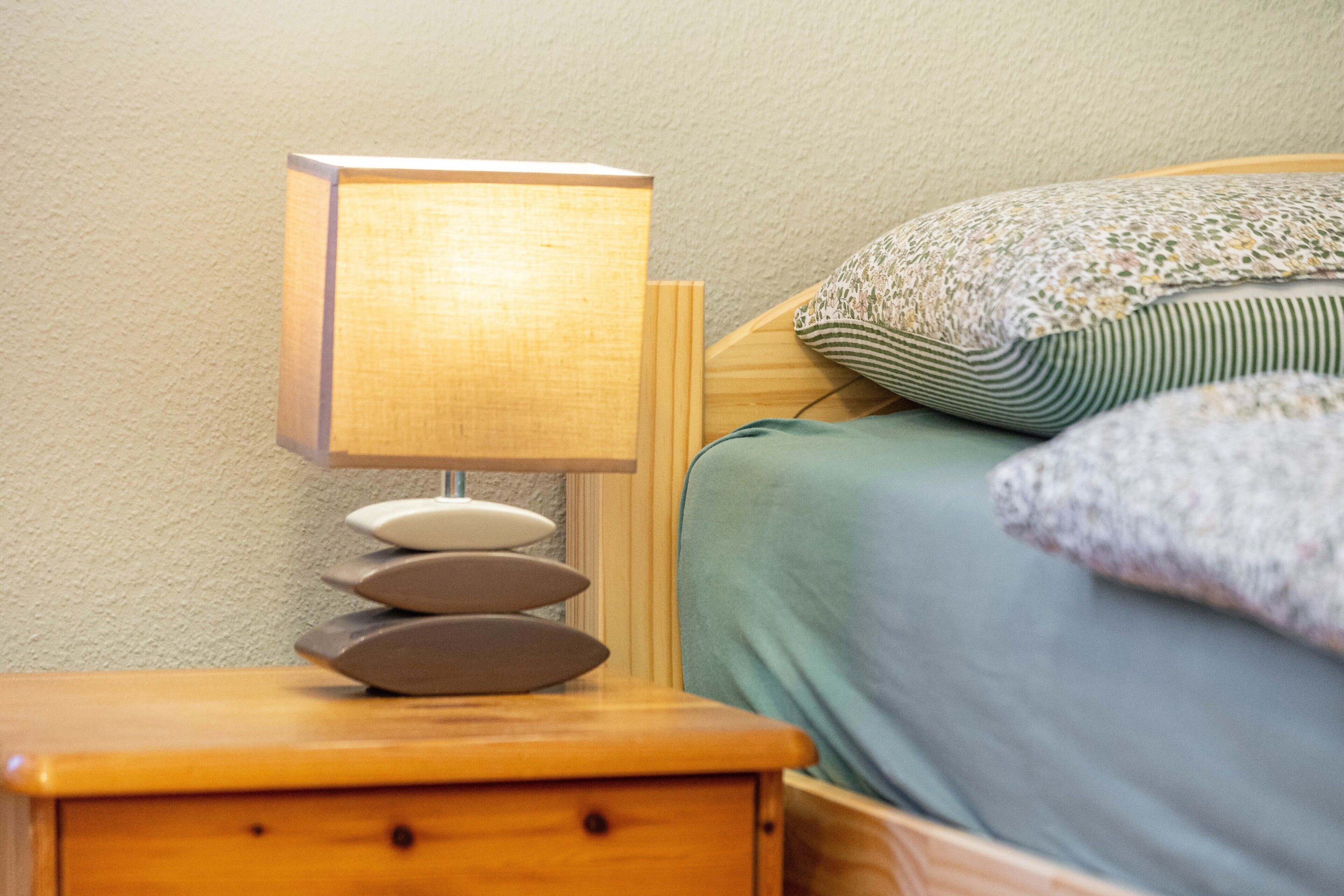 Una mesita de noche de madera con una lámpara, una cama con sábana verde y varias almohadas.