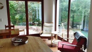 Una habitación con una mesa de comedor de madera, un sillón blanco, una silla roja y puertas correderas de cristal que dan a un jardín.