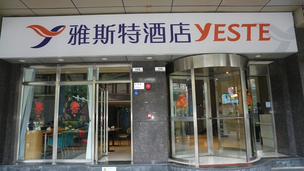 L’entrée d’un hôtel, avec des portes en verre, une enseigne au-dessus indiquant « YESTE », et un écran numérique affichant les disponibilités des chambres.