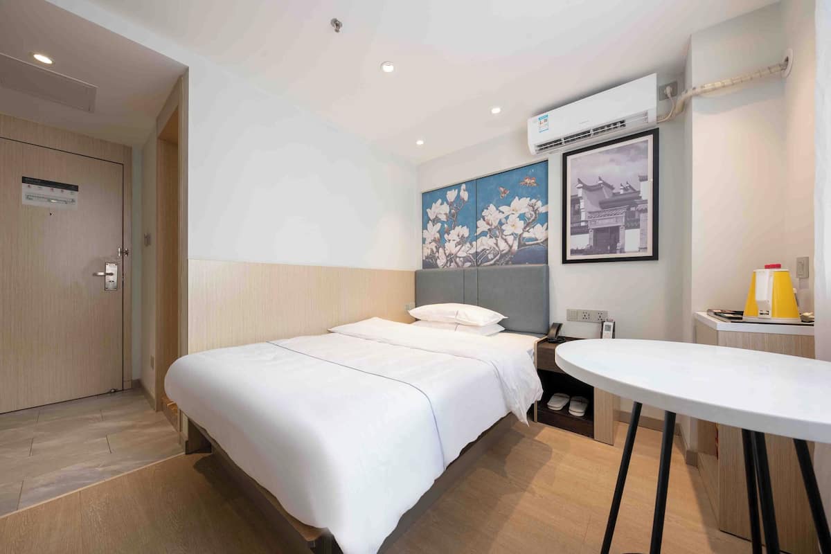 A hotel room with a bed, a small table, a nightstand, and a wall-mounted air conditioner.