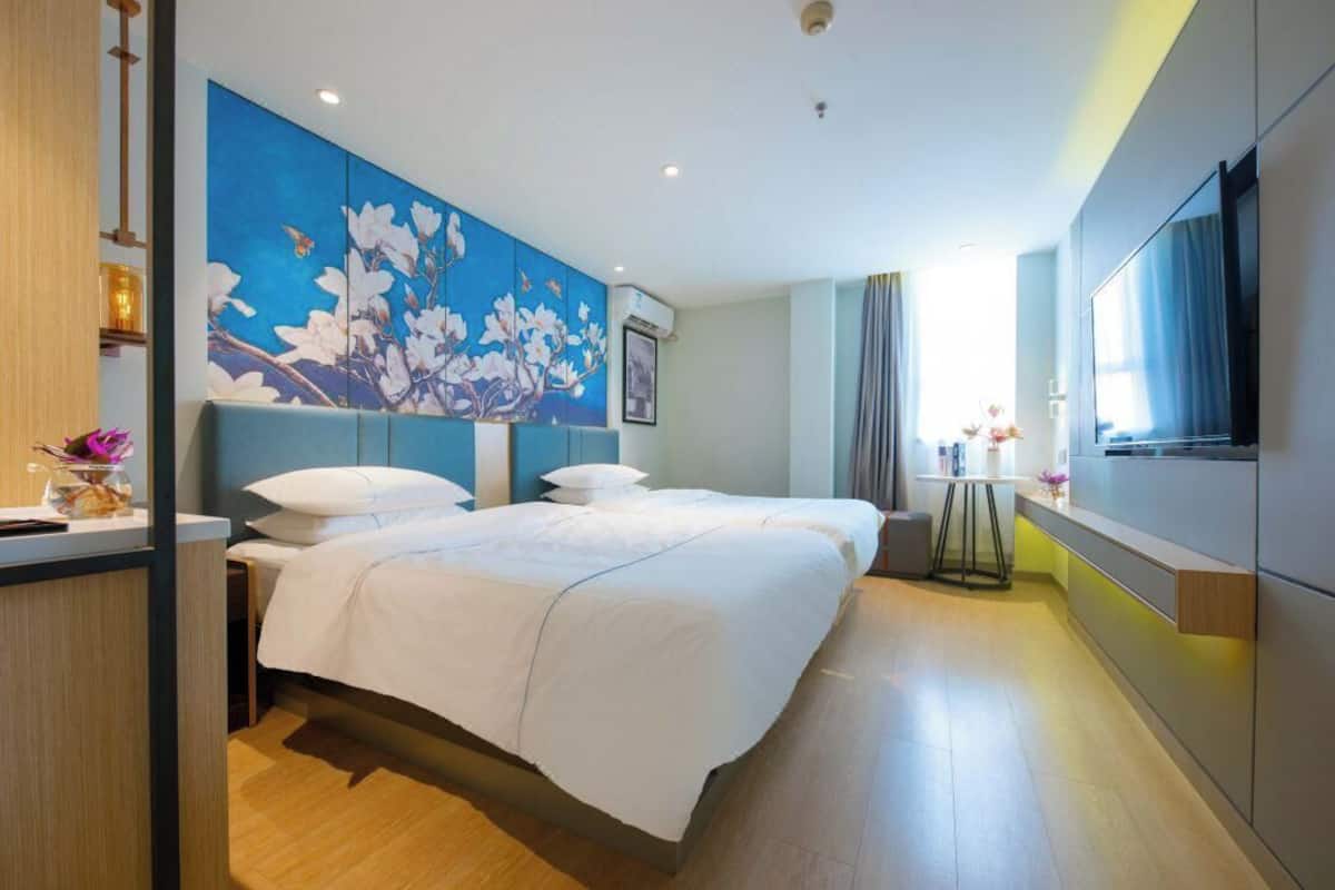 A hotel room with a large bed, a TV, a painting on the wall, and a small table with a vase.