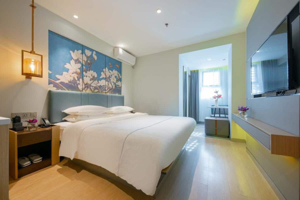 A modern hotel room with a large bed, a bedside table with a lamp, a painting on the wall, and a TV mounted on the wall.
