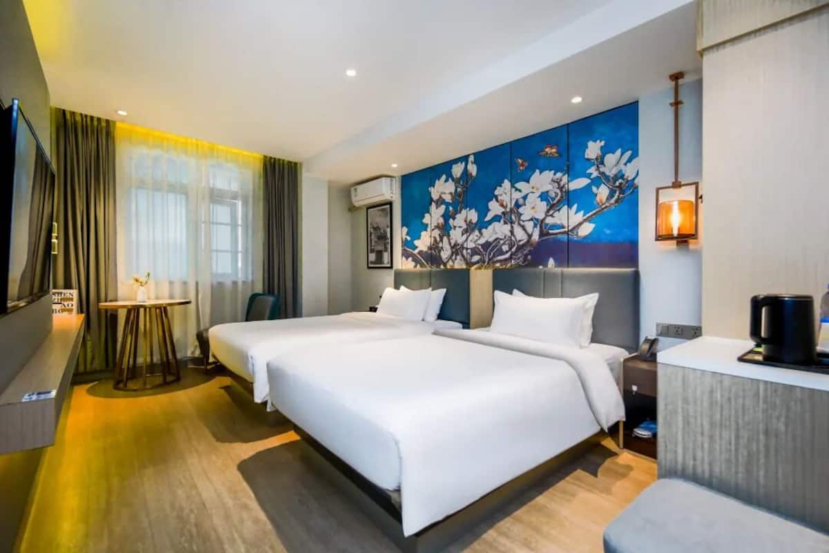 A hotel room with two beds, a desk, a chair, a painting, and a lamp.