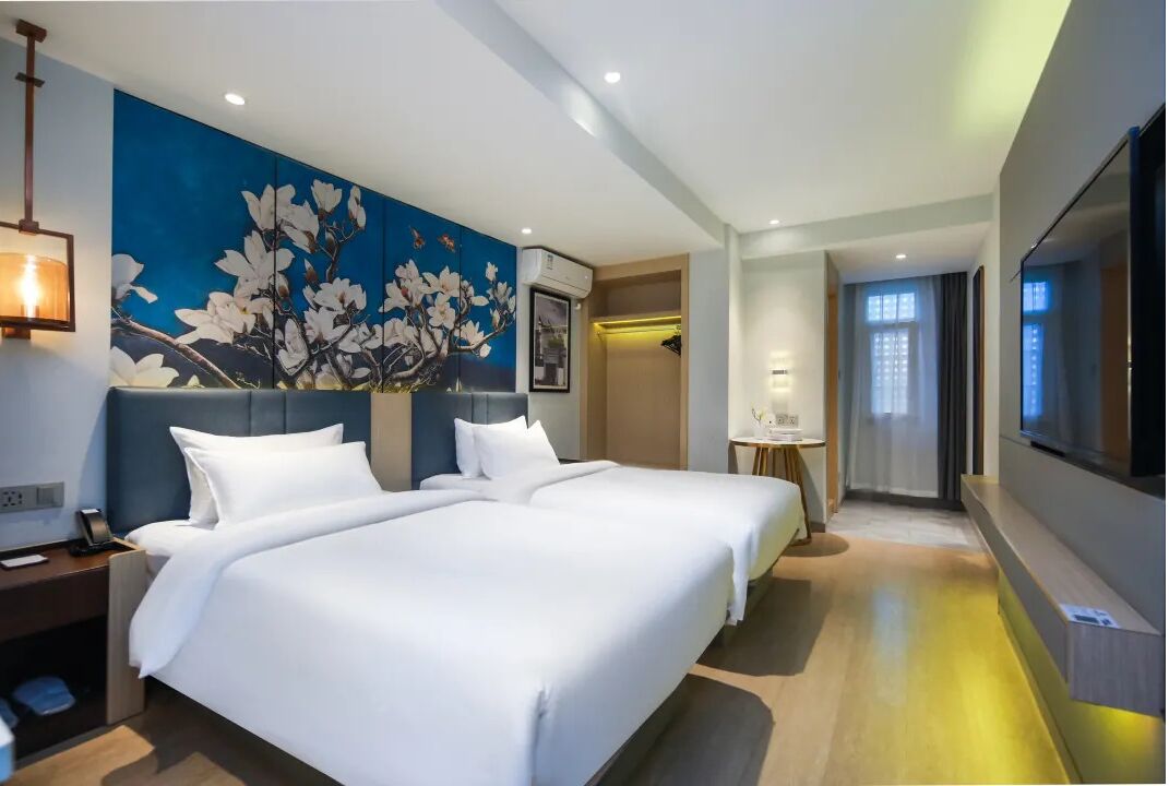 A hotel room with two beds, a large floral mural on the wall, a flat-screen TV, and a small table with a lamp.